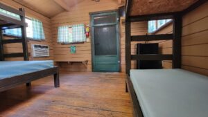 A simple wooden cabin interior with two bunk beds, green checkered curtains, an air conditioner, a fire extinguisher, a small bench, and a dark green door on a wooden floor.