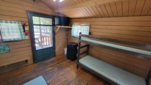 Interior of a wooden cabin with bunk beds, a small fridge, a wall-mounted TV, two windows with green-checked curtains, a door with glass panes, and hardwood floors. Sunlight filters in from outside.