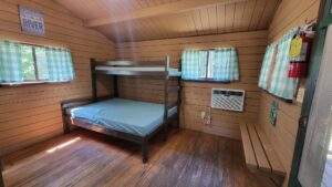 A small wooden cabin room with a bunk bed, green plaid curtains on the windows, an air conditioner, a fire extinguisher, and a wooden bench. Sunlight filters in, creating a cozy and rustic atmosphere.