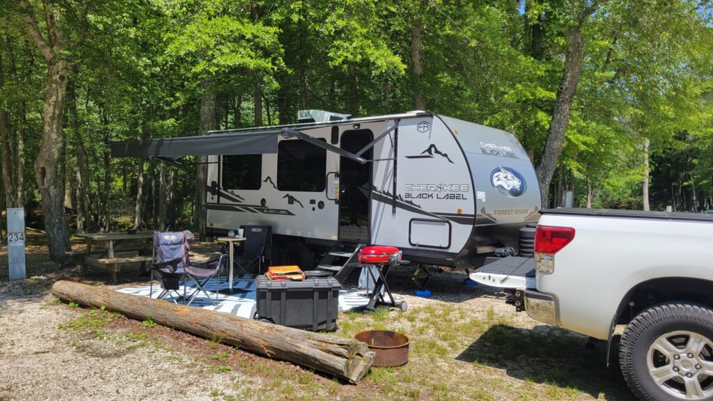 A travel trailer camper parked at a wooded campsite with an awning extended. Camping chairs, a table, a cooler, and a grill are set up outside. A white pickup truck is parked nearby, and sunlight filters through the trees.
