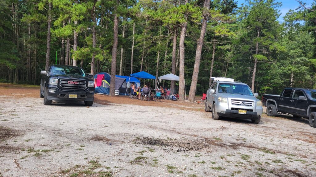 A group of people sits in folding chairs under canopies and near tents at a wooded Grove Group Camping site. Three parked SUVs rest on sandy ground, surrounded by tall pine trees.