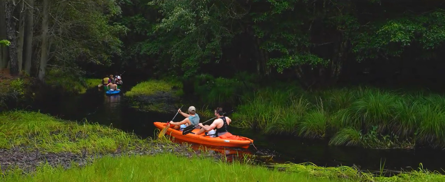 Two people paddle an orange kayak down a narrow, tree-lined river, followed by another group in blue and yellow kayaks. Dense green vegetation surrounds the water.