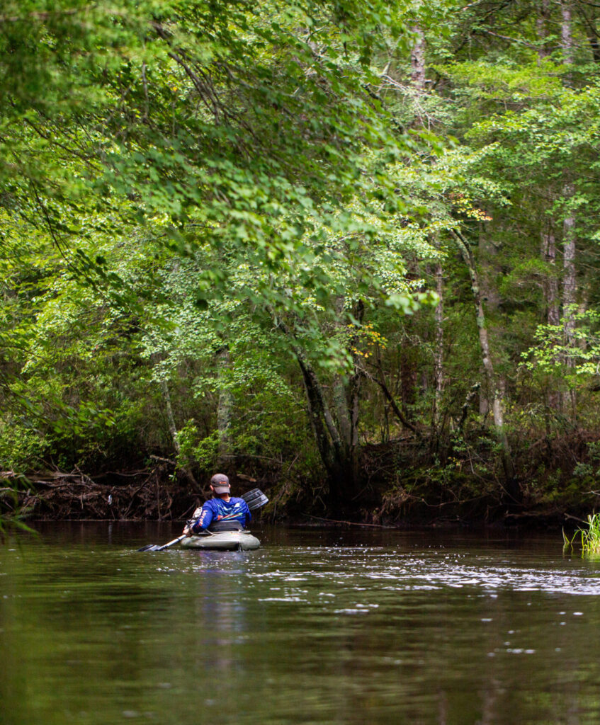 A person paddles an inflatable raft down a calm, tree-lined river surrounded by dense green forest.