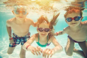 Three children with goggles swim underwater in a pool, looking at the camera with sunlight filtering through the water.