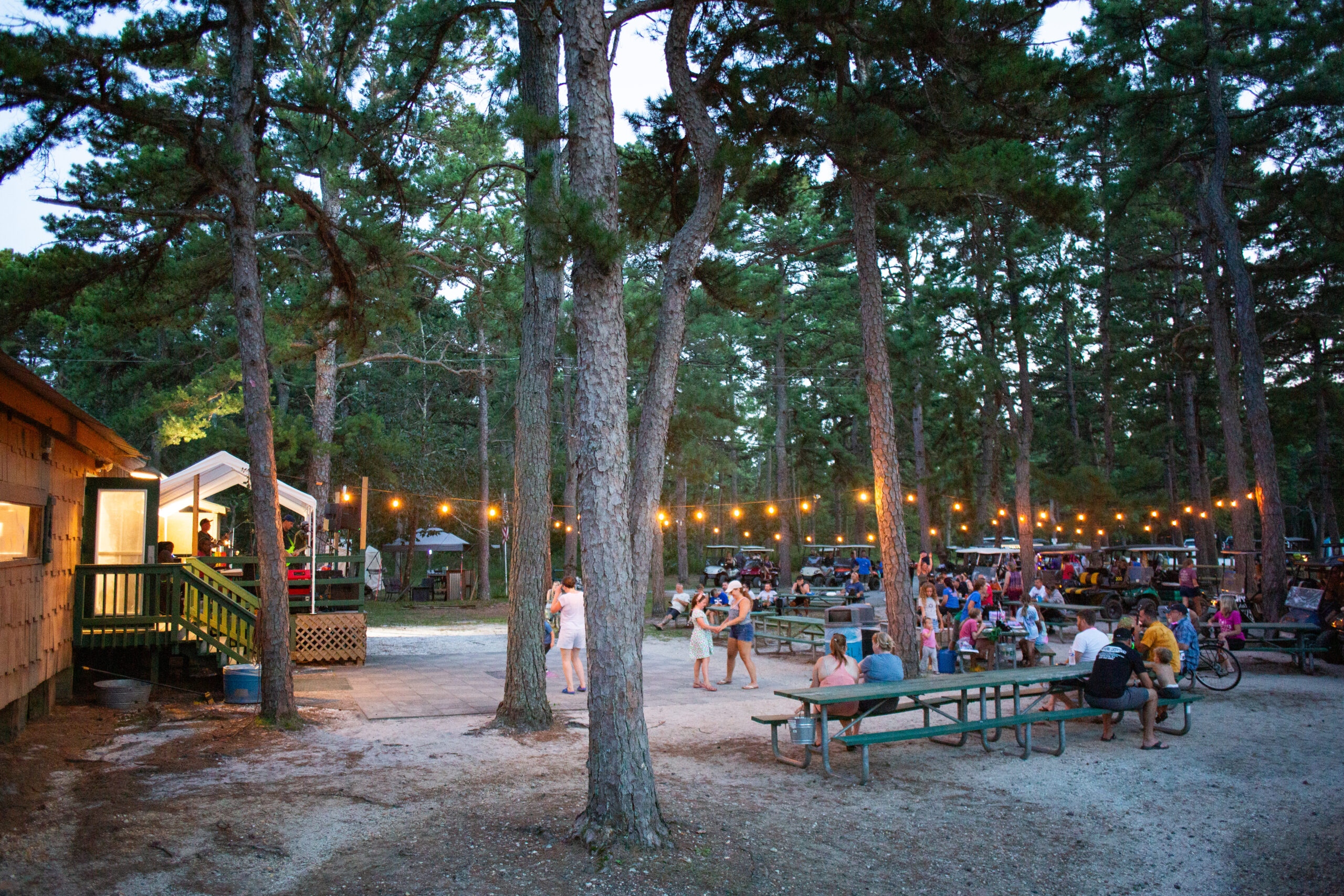 People gather at picnic tables under string lights in a wooded outdoor area; some stand, others sit, with buildings and trees surrounding the space.