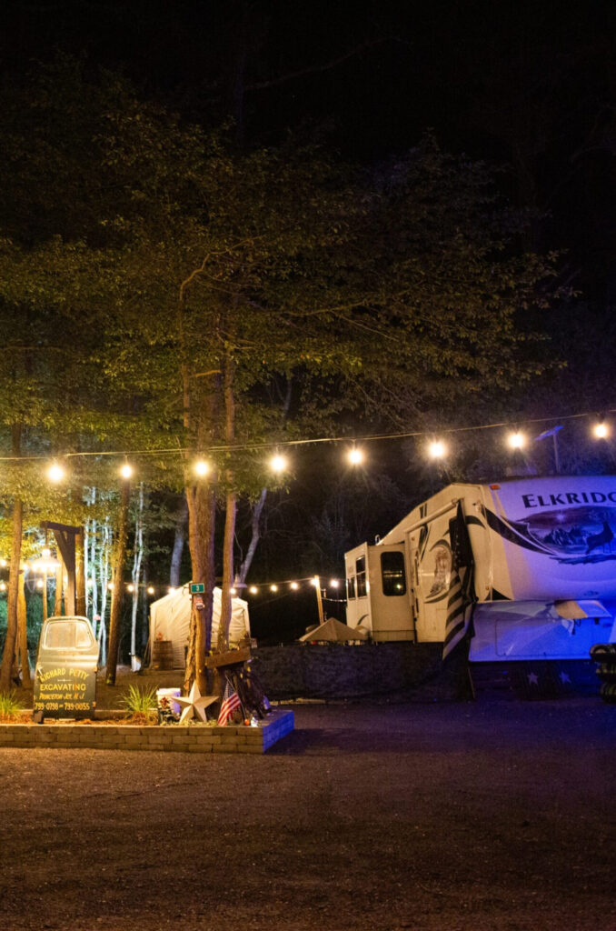 String lights hang above a campsite at night, with an RV and a small tent visible among tall trees. A lit sign stands near a picnic area in the foreground.