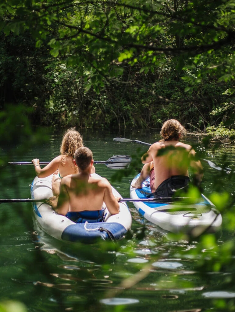 Three people paddle inflatable kayaks on a calm river surrounded by green foliage, viewed from behind through tree branches.