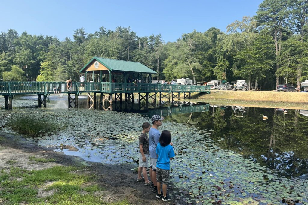 Three children stand near the edge of a lily pad-covered pond, with a wooden dock, pavilion, and parked vehicles visible in the background.