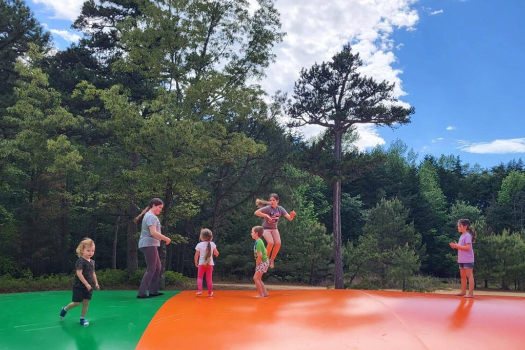 Several children and an adult play and jump on a large outdoor inflatable pad, surrounded by trees and under a partly cloudy sky.