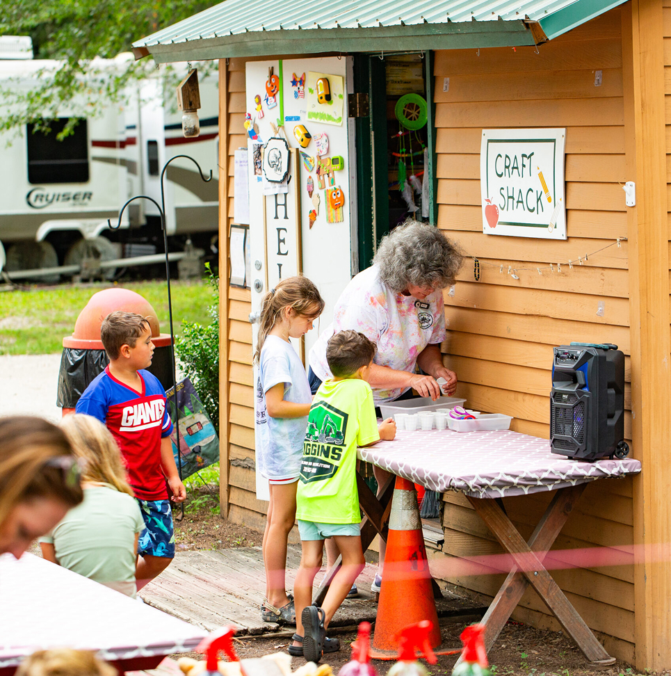 An adult helps three children with a craft activity at an outdoor table beside a small wooden building labeled "Craft Shack." Other people and a camper are visible in the background.