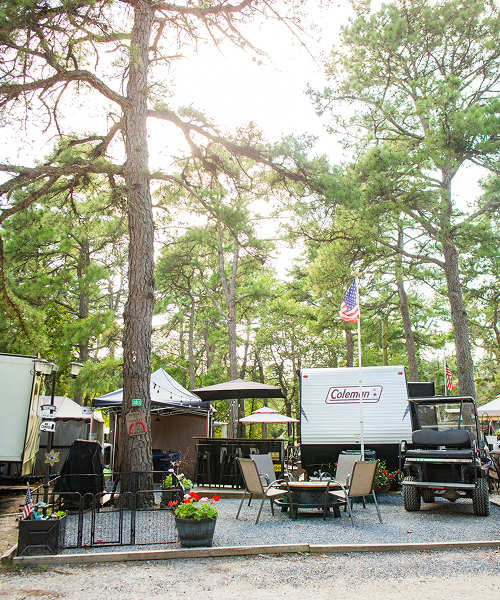A campsite with a Coleman RV, outdoor seating area, grill, potted flowers, and an American flag, surrounded by tall pine trees in daylight.