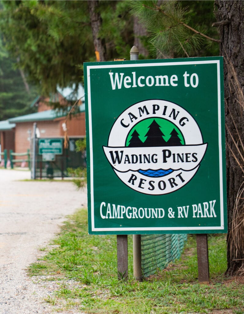 Green sign reads "Welcome to Camping Wading Pines Resort Campground & RV Park" with trees and a building visible in the background.