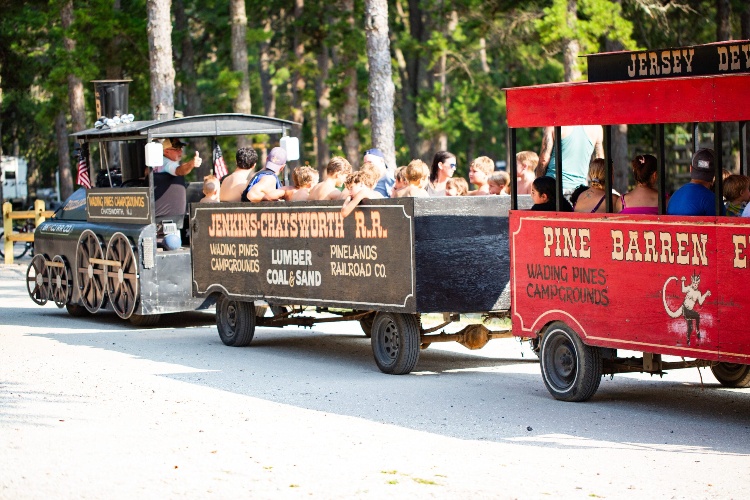 A small open-air train with passengers sits on a path in a wooded area. The train cars display signs for the Jersey-Shatsworth Railroad and Pine Barrens Campgrounds.