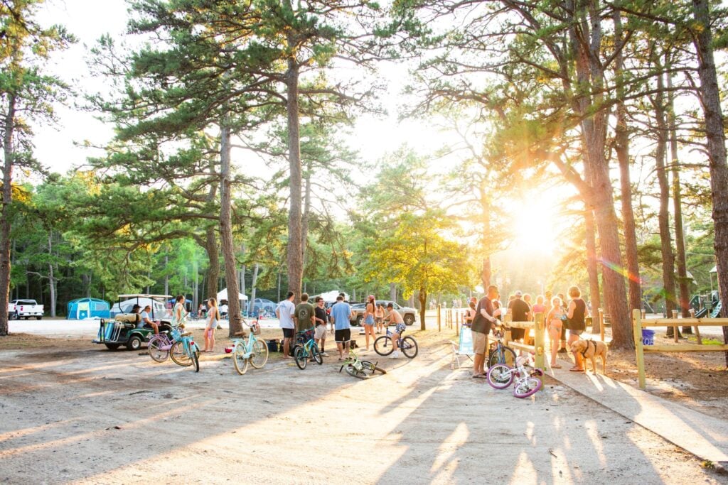 A group of people gathers near bicycles in a sunlit forest campground, with tents and vehicles visible in the background.