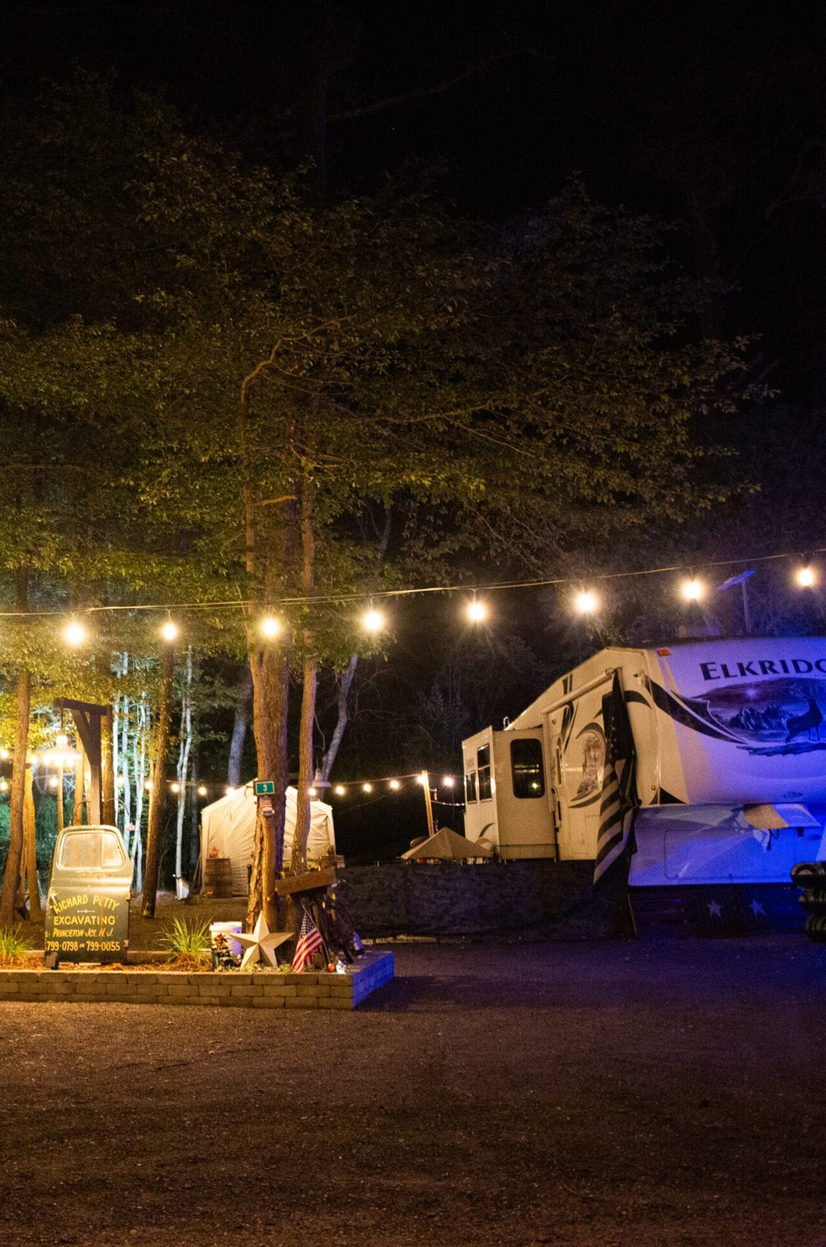 String lights hang above a campsite at night with an RV, picnic table, chairs, and a sign reading "Camp Here Incognito." Trees and parked vehicles are visible in the background.