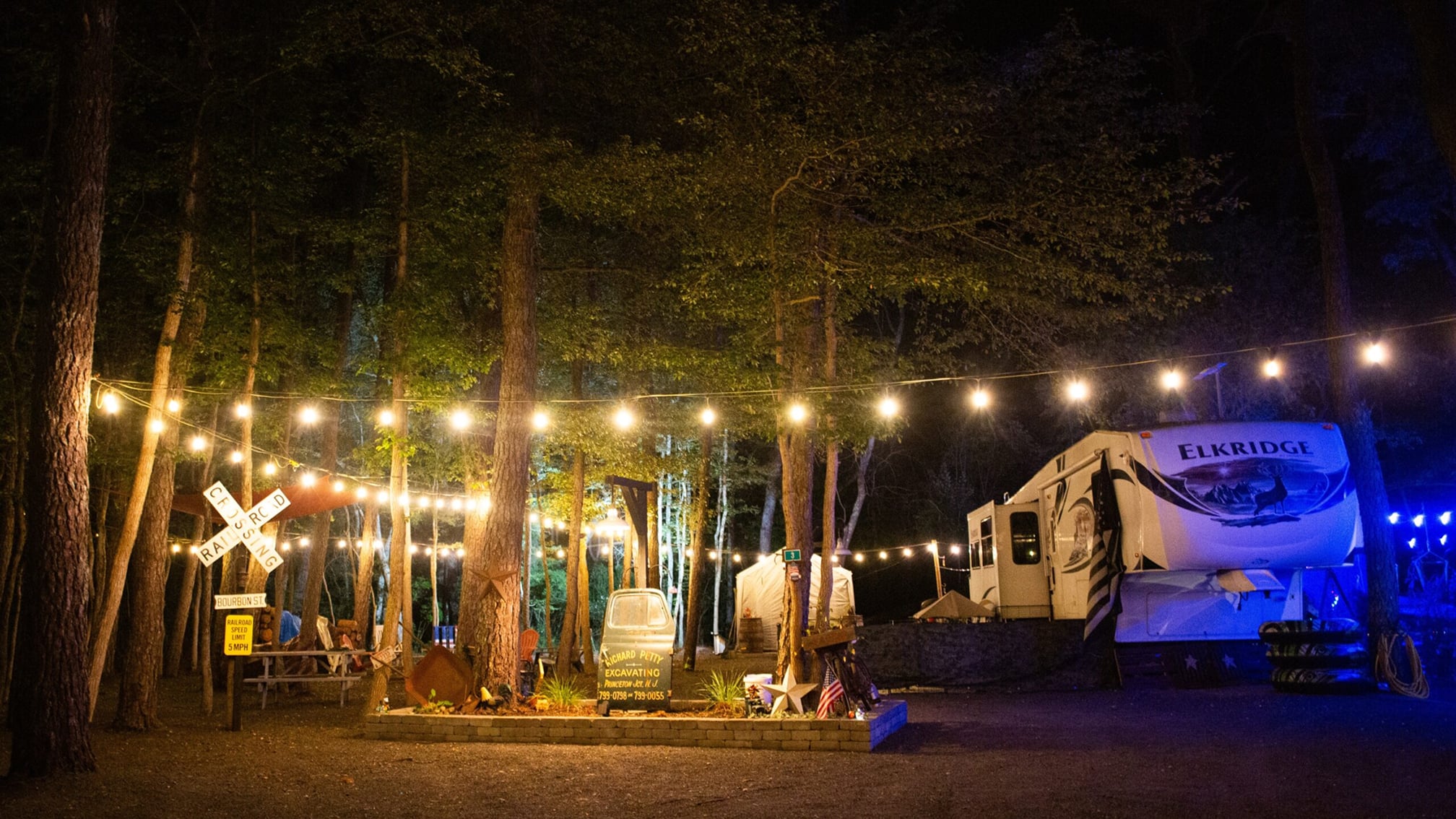 A campground at night with string lights overhead, an RV labeled “Elkridge,” a picnic table, railroad crossing signs, and trees surrounding the area.