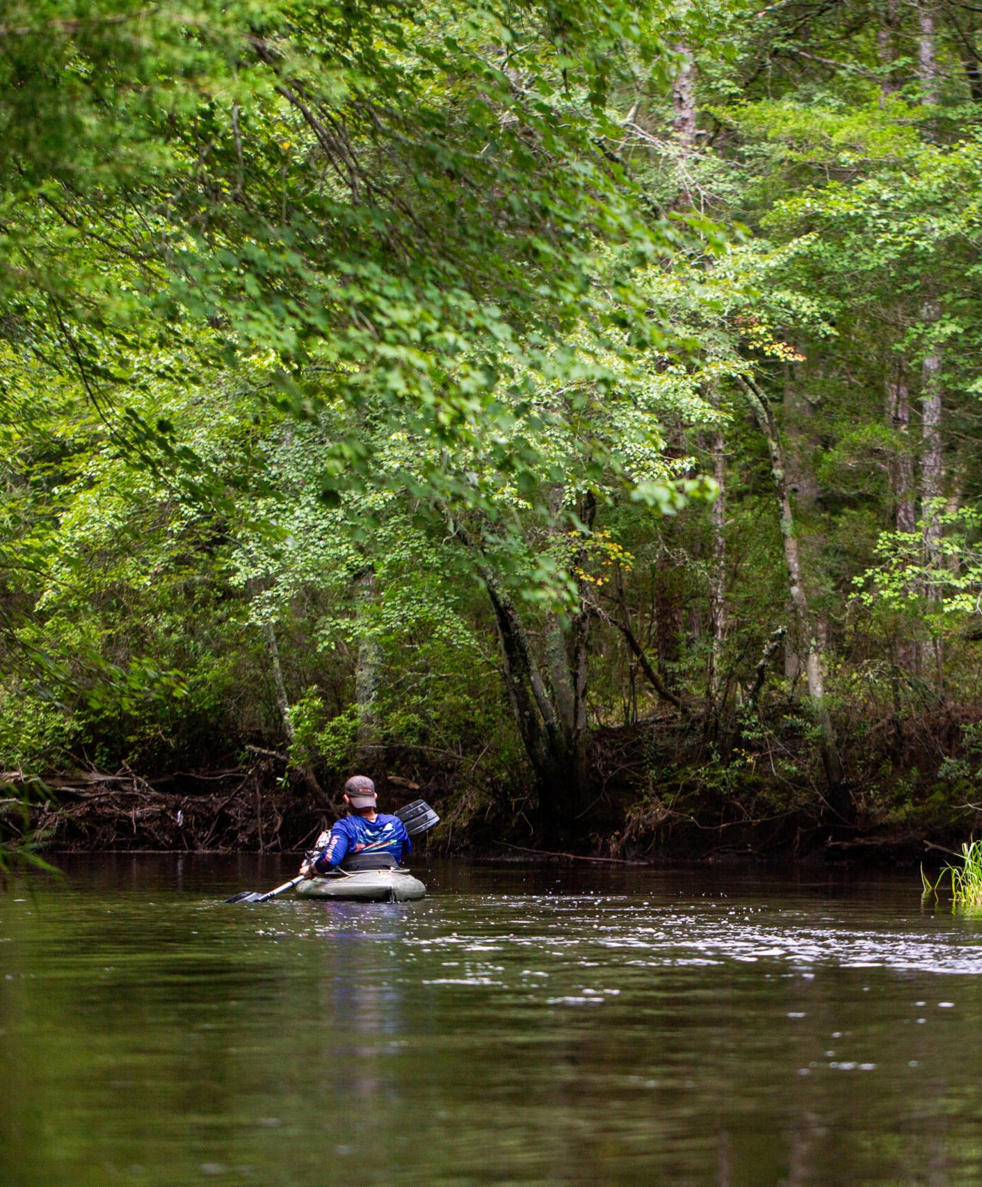 A person in a blue shirt paddles an inflatable raft down a calm, tree-lined river surrounded by dense green forest.