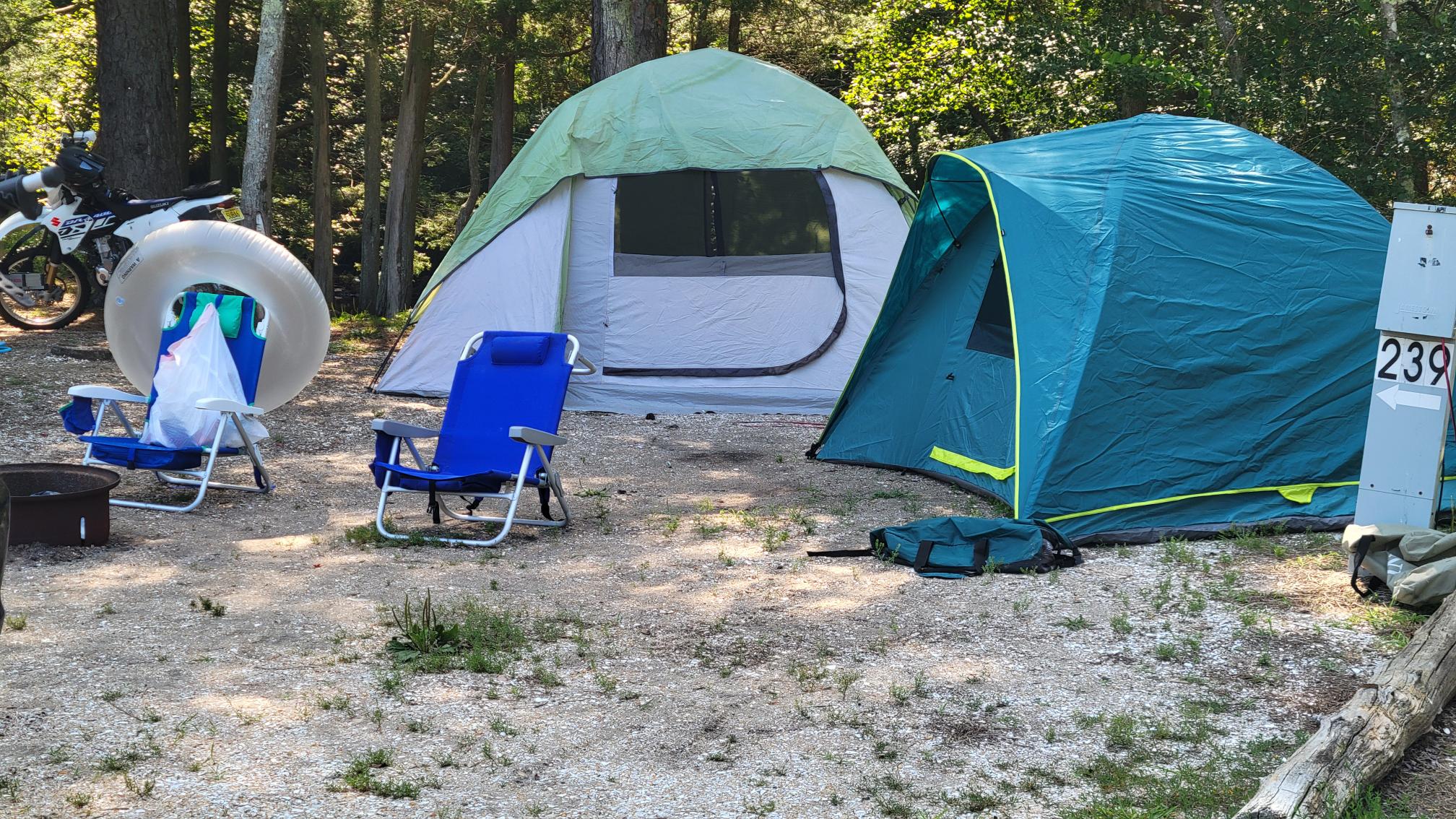 Two tents, two blue chairs, a white inner tube, and a motorcycle are set up at a campsite marked 239 in a wooded area.