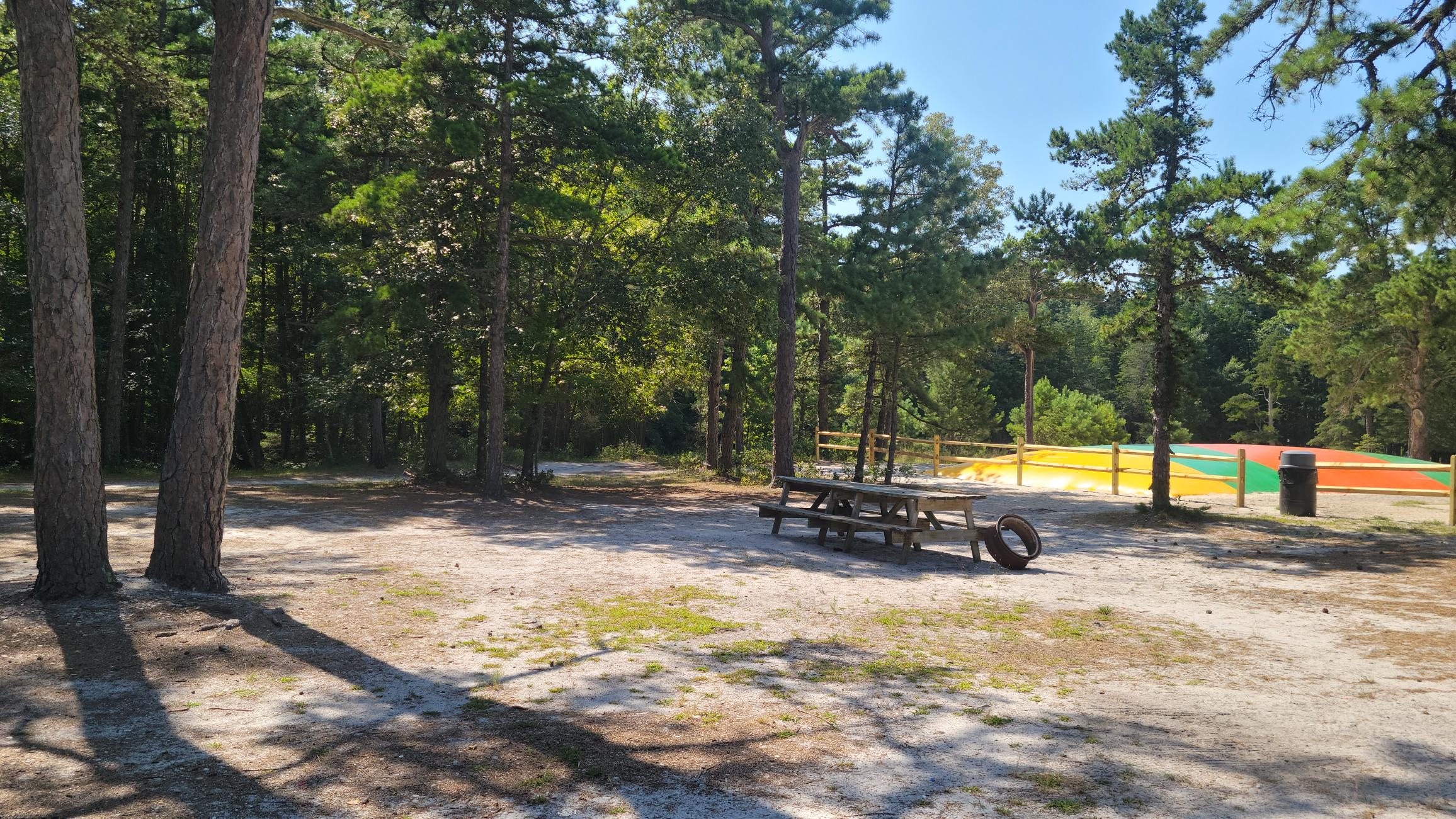 A wooden picnic table and tire sit on sandy ground among trees near a fenced colorful inflatable attraction in a sunny outdoor setting.