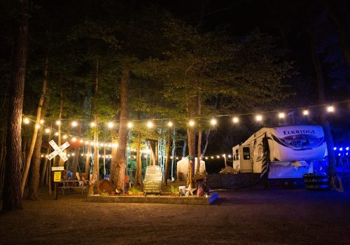 A well-lit campsite at night with string lights, an RV labeled "Elkridge," tents, picnic tables, and trees in the background.