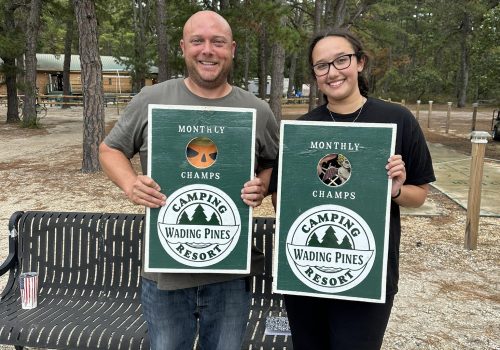 Two people stand outdoors holding "Monthly Champs Camping Wading Pines Resort" signs, smiling in front of a bench and trees.